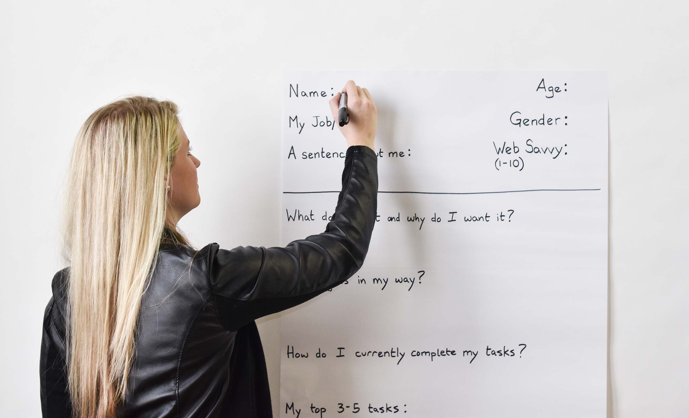 A blonde woman writes on a user persona worksheet that is affixed to a wall.